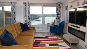 A living room with a sofa, a blue armchair, a TV, and a view of the sea. - Bay View, Tresaith - Two Bedroom Cottage, Sleeps 4 (Tresaith)