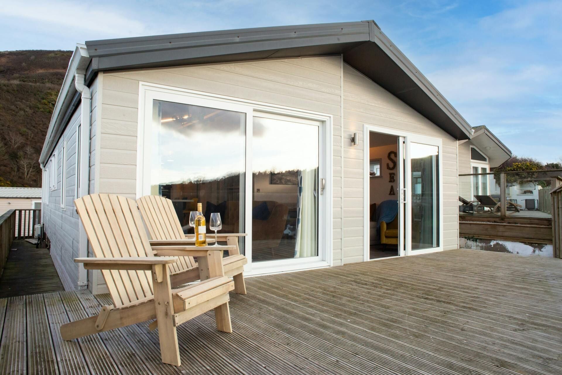 A wooden deck with two chairs and a table, a sliding glass door to a room, and a view of the surrounding landscape.