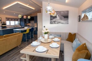 A modern kitchen with blue cabinets, a dining area with a wooden table set for four, and a living area with a sofa and cushions. - Bay View, Tresaith - Two Bedroom Cottage, Sleeps 4 (Tresaith)