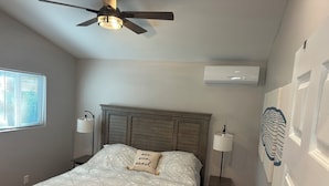 Un dormitorio con cabecera de madera, cama con ropa de cama estampada, dos lámparas de noche, ventilador de techo, ventana con persianas y aire acondicionado de pared.