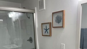 Un baño con cabina de ducha de vidrio, un inodoro blanco, un lavabo con espejo y dos cuadros enmarcados en la pared.