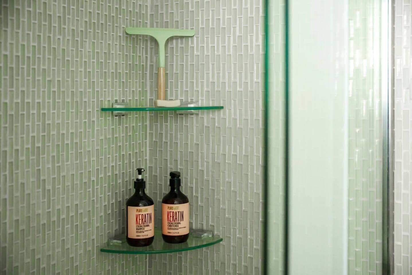A bathroom with a patterned wall, a green cleaning tool, and two bottles of Pure Keratin shampoo on a glass shelf.