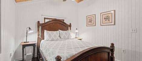A bedroom with a wooden bed, two bedside lamps, a ceiling fan, and two framed pictures on the wall.