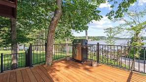 A wooden deck with a barbecue grill, surrounded by a metal railing, overlooking a body of water and greenery.