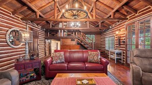 A log cabin interior with a leather sofa, wooden staircase, and a large wooden wheel chandelier.