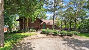 A log cabin with a gravel driveway leading to a green door, surrounded by trees and a fenced yard.