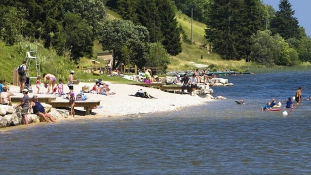 A lakeside beach with people sunbathing and swimming, surrounded by dense forest. - Cosy duplex in the Haut-Jura with detached garage. (PREMANON)