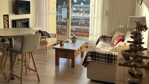 A spacious living area with a wooden ceiling, a dining table with chairs, a sofa with a plaid blanket, and a view of a traditional building through large windows. - Cosy duplex in the Haut-Jura with detached garage. (PREMANON)