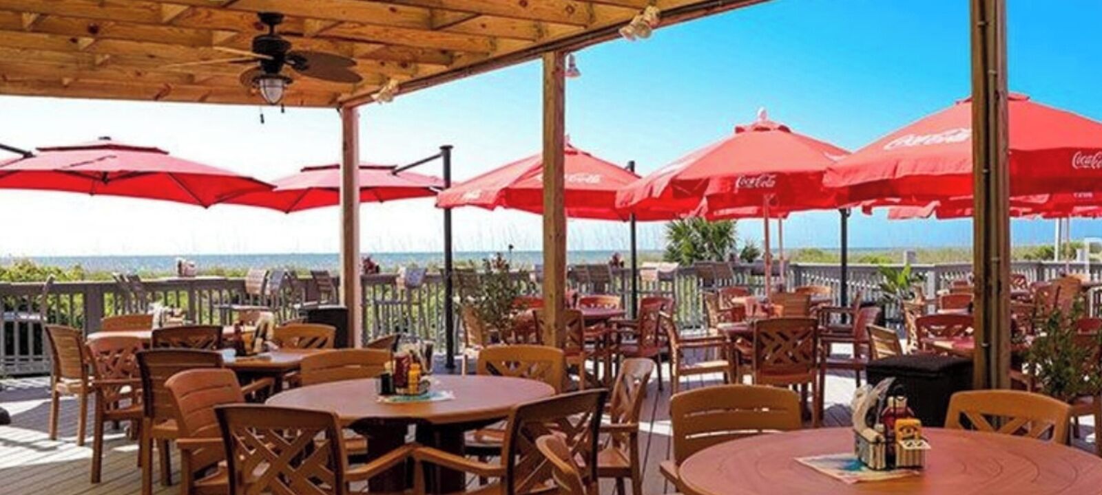 An outdoor dining area with wooden tables and chairs, red umbrellas, and a view of the ocean.