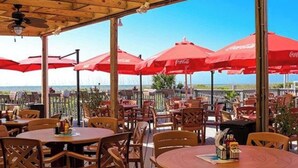 Un espace extérieur aménagé pour les repas, avec des tables et des chaises en bois, des parasols rouges et une vue sur l’océan.