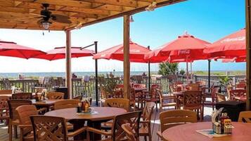 An outdoor dining area with wooden tables and chairs, red umbrellas, and a view of the ocean.