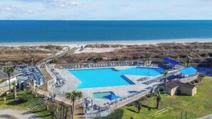 An aerial view of a resort with a large central pool, a beach, and a fenced area with a small building.