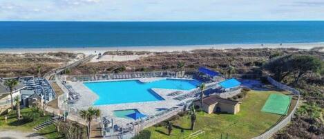 An aerial view of a resort with a large central pool, a beach, and a fenced area with a small building.