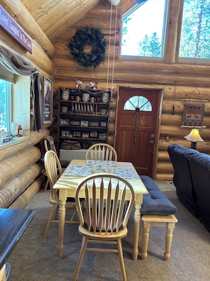 Un interior de cabaña acogedor con una mesa de madera dispuesta para cuatro personas, rodeada de sillas, una estantería con varios objetos y una puerta con un ventanal circular.