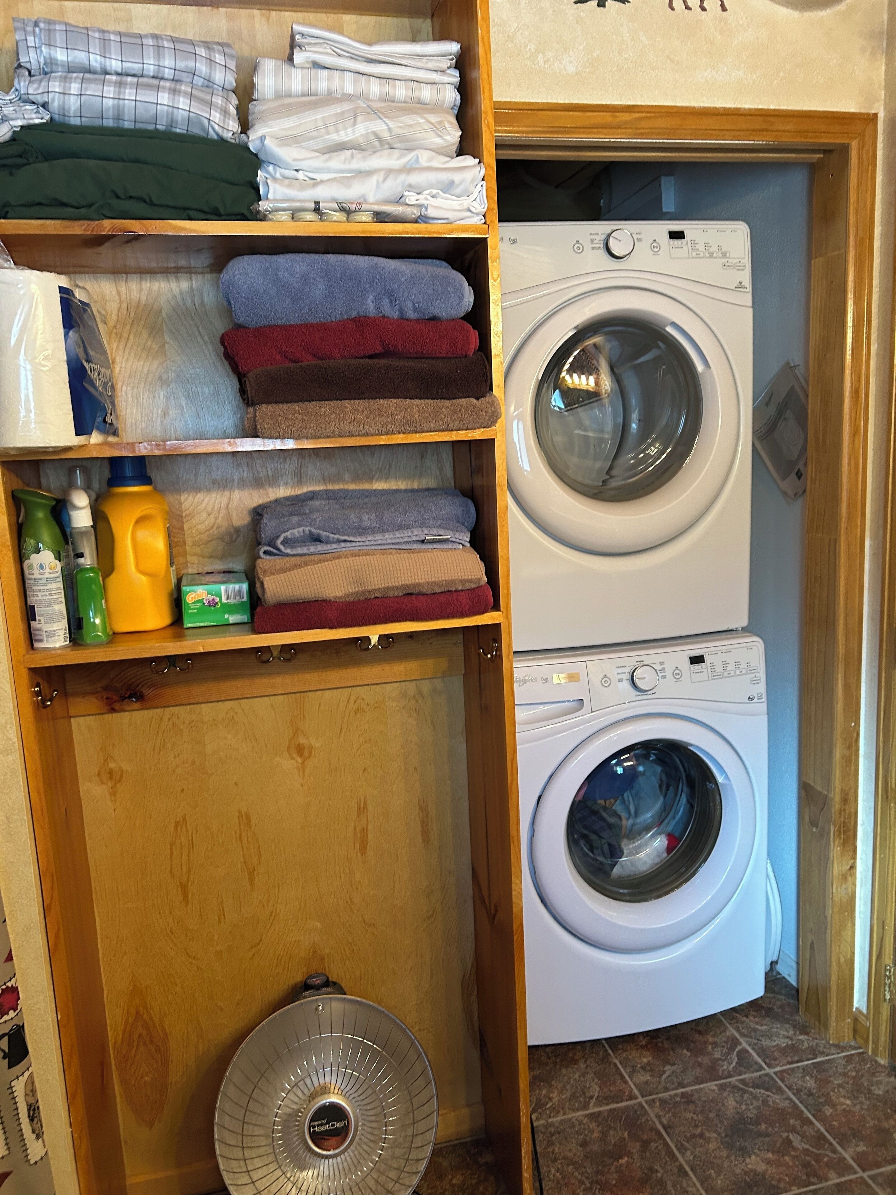 Un espace buanderie comprenant une machine à laver et un sèche-linge superposés, des étagères avec des serviettes pliées et des produits de nettoyage, et un ventilateur.