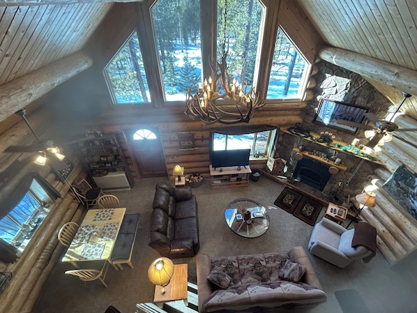 Interior de una cabaña de troncos con chimenea, muebles de madera y amplias ventanas que ofrecen vistas al bosque.
