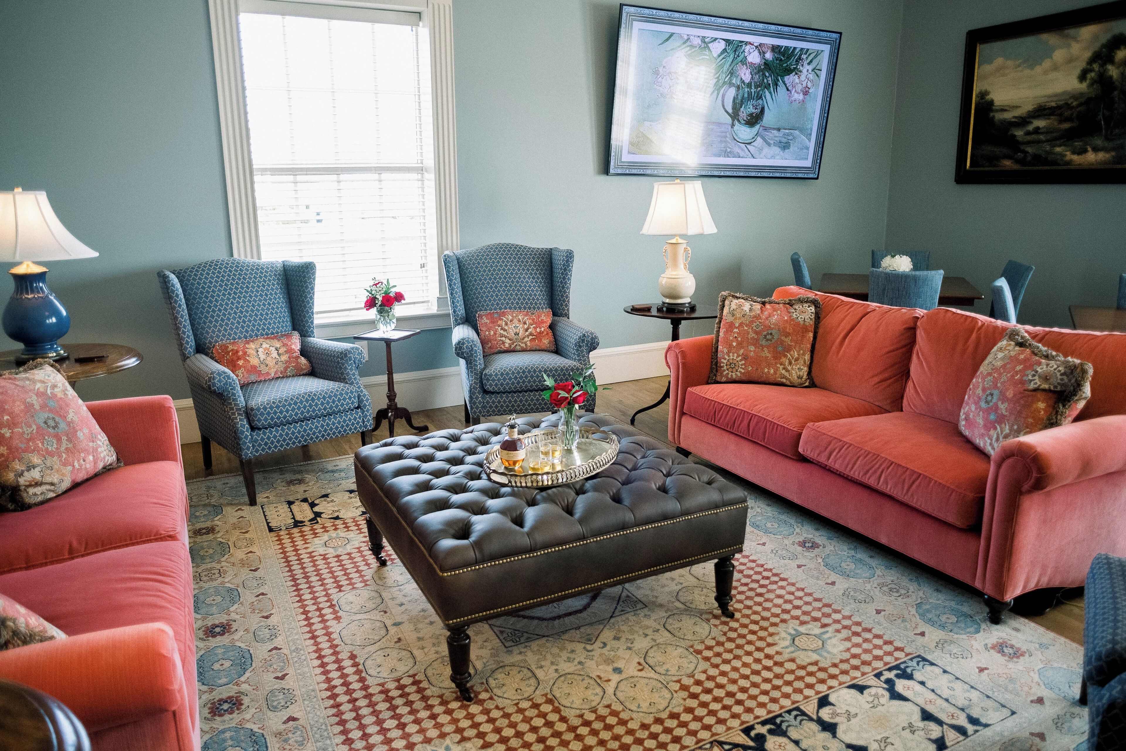 Un salon avec un canapé rouge, des fauteuils bleus, un pouf marron et des tapis à motifs.