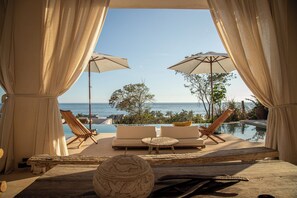 Un área de descanso junto a la piscina con vista al océano, que incluye sillones reclinables, sombrillas y una mesa de madera.