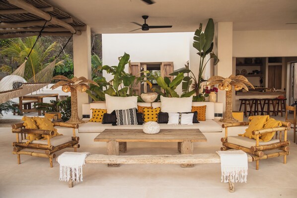 A covered outdoor seating area with a wooden table, cushioned chairs, and large potted plants.
