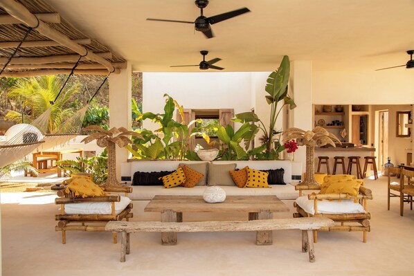 A spacious living area with a wooden sofa, two wooden benches, and a central coffee table. There are ceiling fans, a hanging hammock, and a variety of plants.