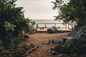 Une terrasse en bois avec un banc donnant sur une plage de sable, entourée de rochers et de végétation.