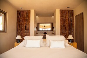 A double bed with white linens, two bedside tables with lamps, a large mirror above the bed, and a view of the bathroom through a doorway. - Casa Selva, Troncones (Troncones)