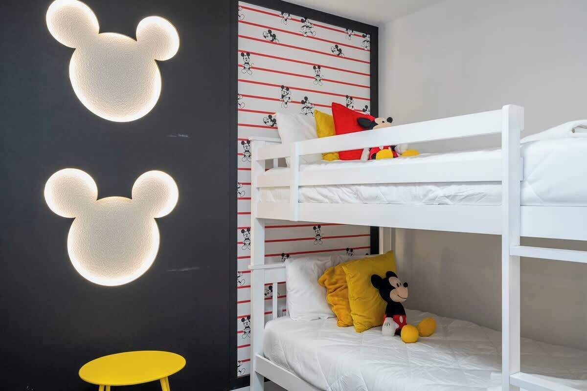 Una cama individual con decoración temática de Mickey Mouse, una mesita amarilla pequeña y un peluche de Mickey Mouse.