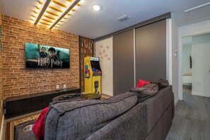 Un salon moderne avec un mur en briques, un téléviseur à écran plat, un canapé et une borne d’arcade pour enfants.
