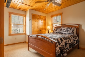 A bedroom with a wooden ceiling, a bed with patterned bedding, and two windows with blinds.