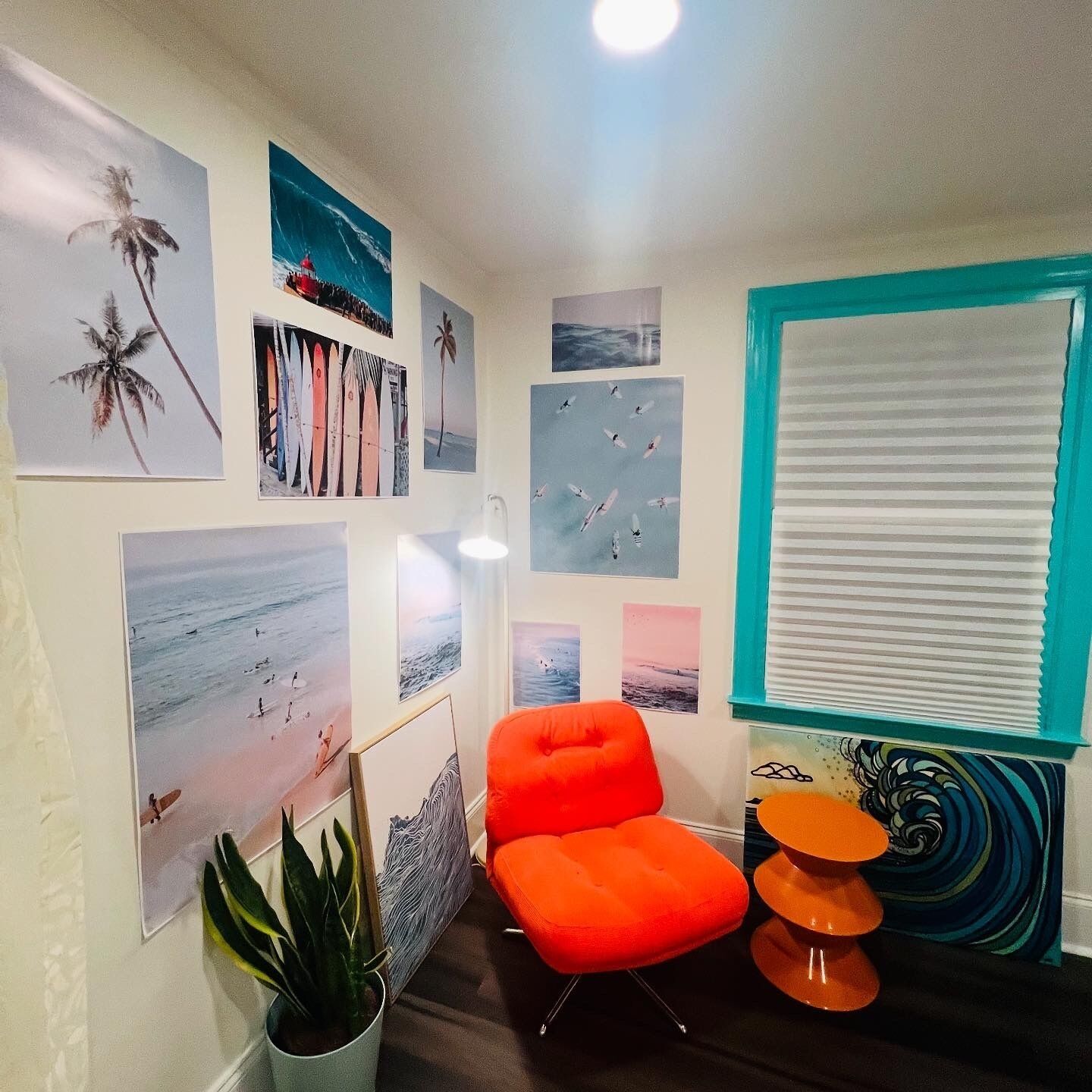 A room with a teal-framed window, a bright orange chair, and multiple beach-themed wall art.