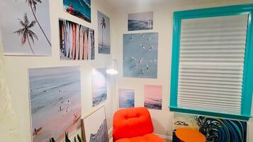 A room with a teal-framed window, a bright orange chair, and multiple beach-themed wall art.
