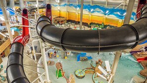 A large indoor water slide with a black tube, surrounded by various play structures and a mural depicting a sunset. - Pocono Lakefront Villa-kayaks, firepl, skiing, gameroom+ (Long Pond)