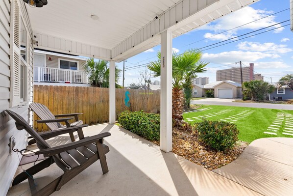 A covered patio with two lounge chairs, a wooden fence, and a well-maintained lawn with a small bush. - Beach House - Sunset by the Sea (Panama City Beach)