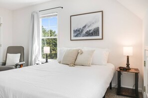 Une chambre d’hôtel avec un grand lit, une chaise, une table de chevet avec une lampe et une fenêtre avec des rideaux.