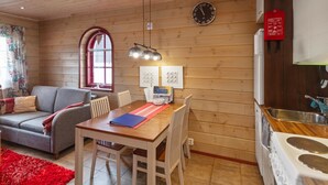 Ein gemütliches Zimmer mit Holzbalkendecke, einem Esstisch mit Stühlen, einer Couch, einem roten Teppich, einer Uhr und einer Küchenzeile mit Herd und Spüle.