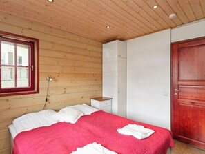 Quarto com teto de madeira, cama com colcha vermelha e branca, mesa de cabeceira branca, guarda-roupa e porta de madeira.
