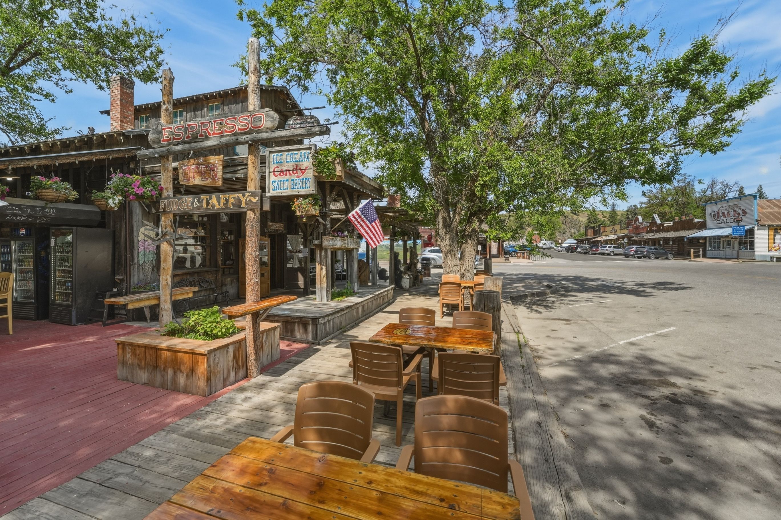 A rustic outdoor seating area with wooden tables and chairs, a storefront with signs for a coffee shop and a sweet bakery, and an American flag.