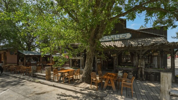 Un espace extérieur aménagé dans un style rustique, avec des tables et des chaises en bois, un grand arbre offrant de l’ombre et un bâtiment sur lequel on peut lire l’inscription « THE RIVER’S ».