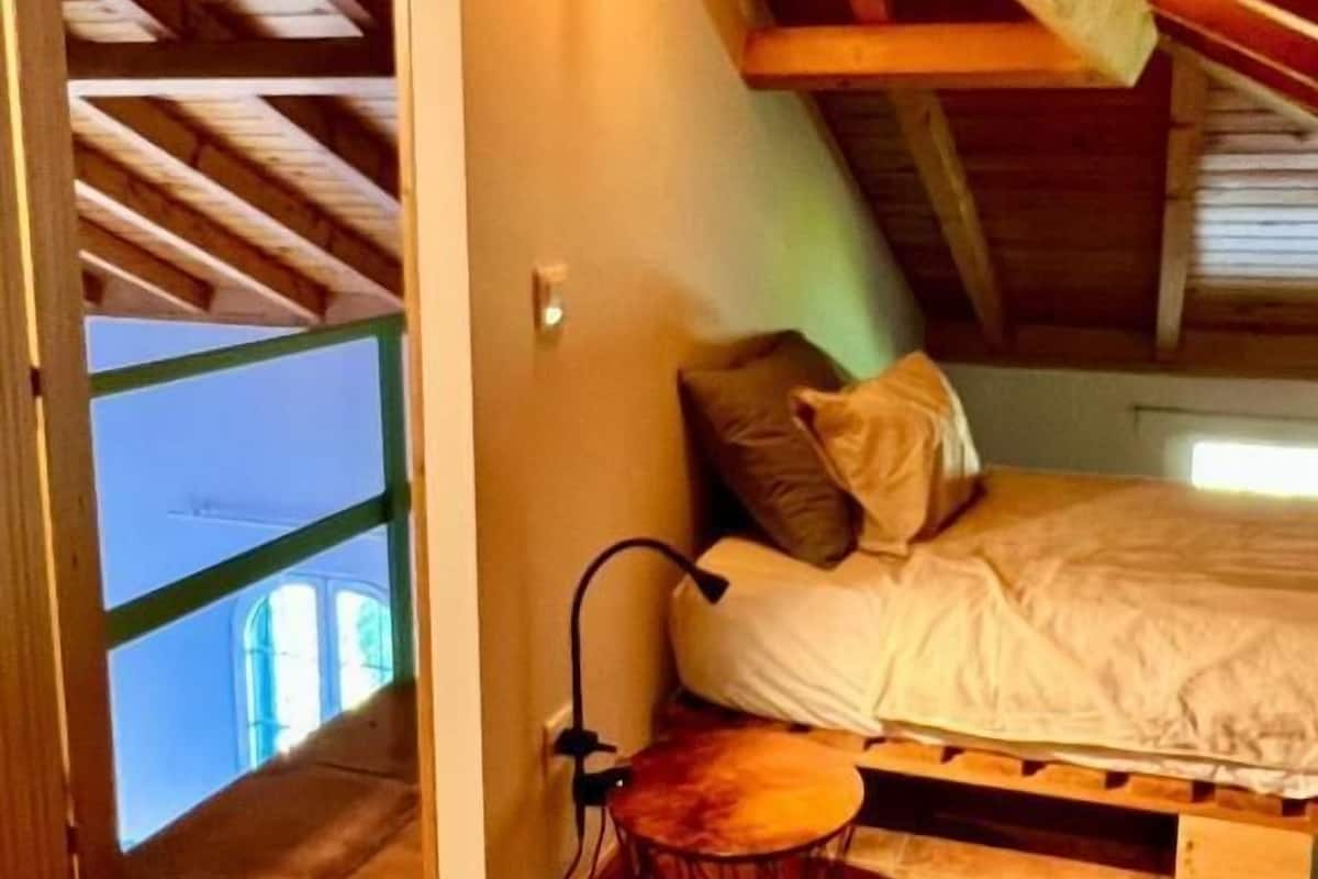 Un dormitorio cálido en el ático con cama, almohadas, una mesita y una silla colgante.