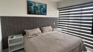 A modern bedroom with a large bed, a nightstand, a painting on the wall, and a window with blinds. - Apartment with comfort, security and exclusivity guaranteed! (Mazatlán)