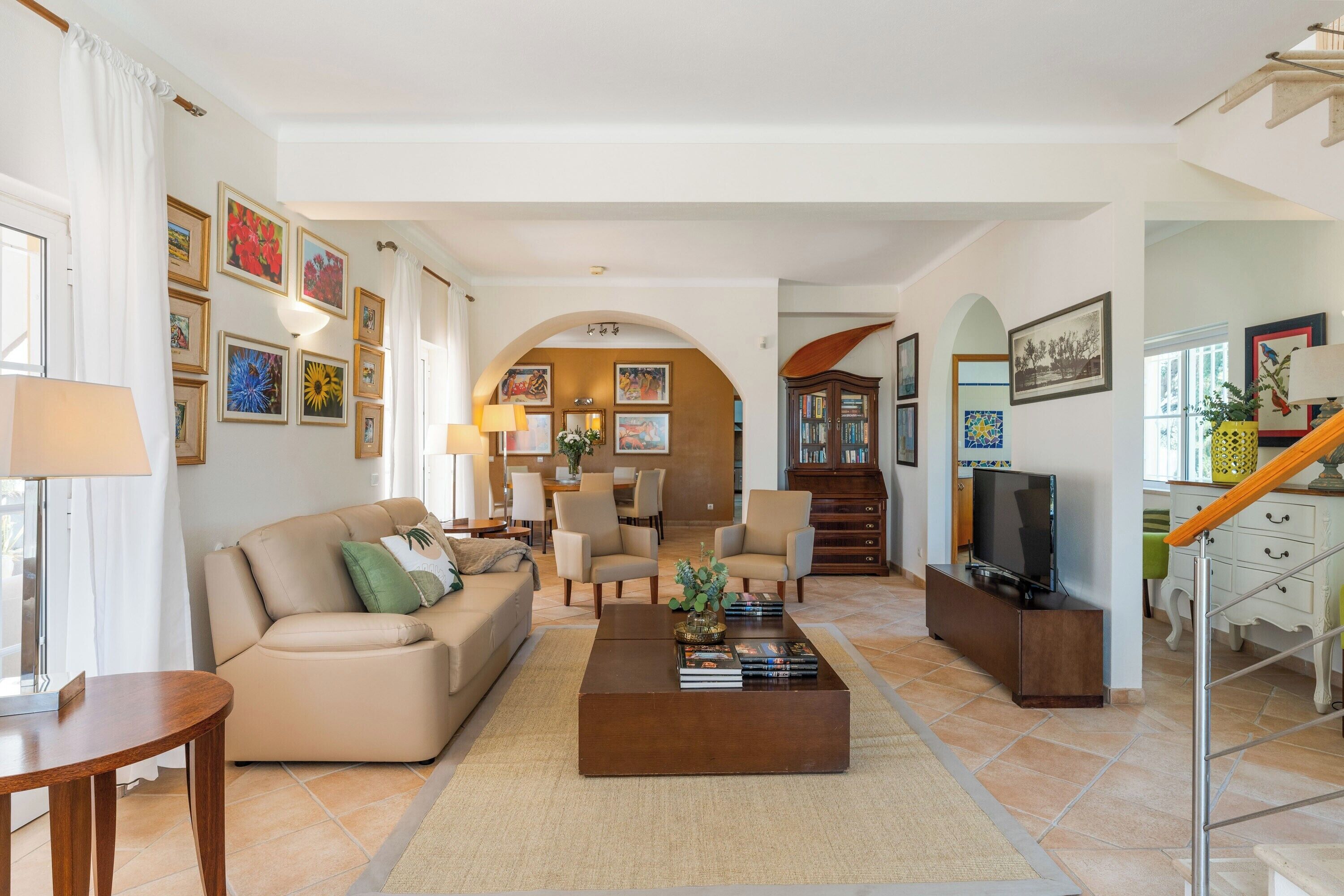 A spacious living room with a sofa, coffee table, and TV. There are framed pictures on the walls and a staircase leading to another level.