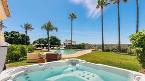 Un jacuzzi dans un jardin, avec une piscine, des palmiers et une maison en arrière-plan.