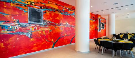 A modern hotel lobby with vibrant abstract wall art, a large white column, and a dining area with round tables.