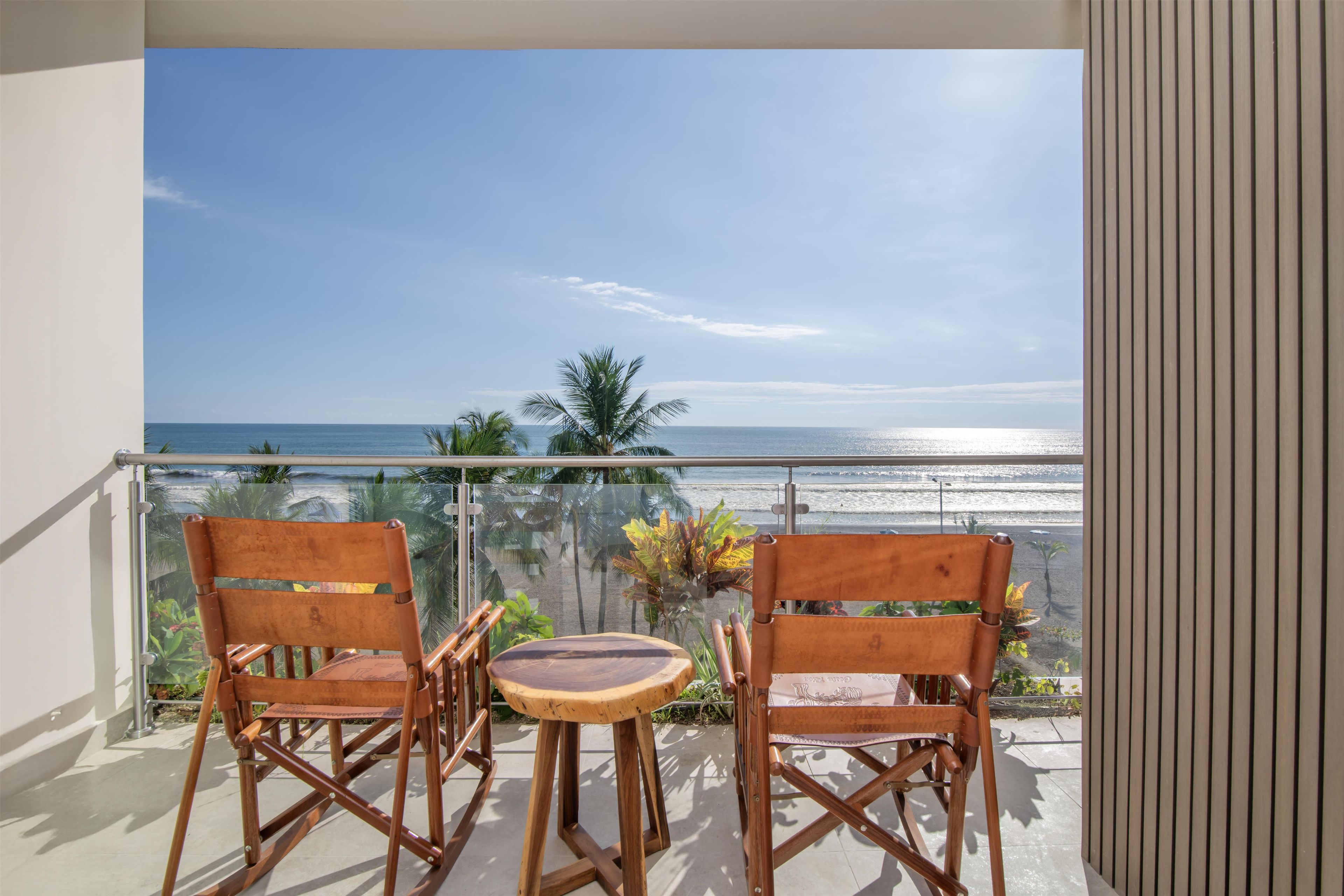Ein Balkon mit zwei Holzstühlen und einem kleinen Tisch, mit Blick auf einen Strand mit Palmen und dem Meer.
