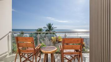 Ein Balkon mit zwei Holzstühlen und einem kleinen Tisch, mit Blick auf einen Strand mit Palmen und dem Meer.