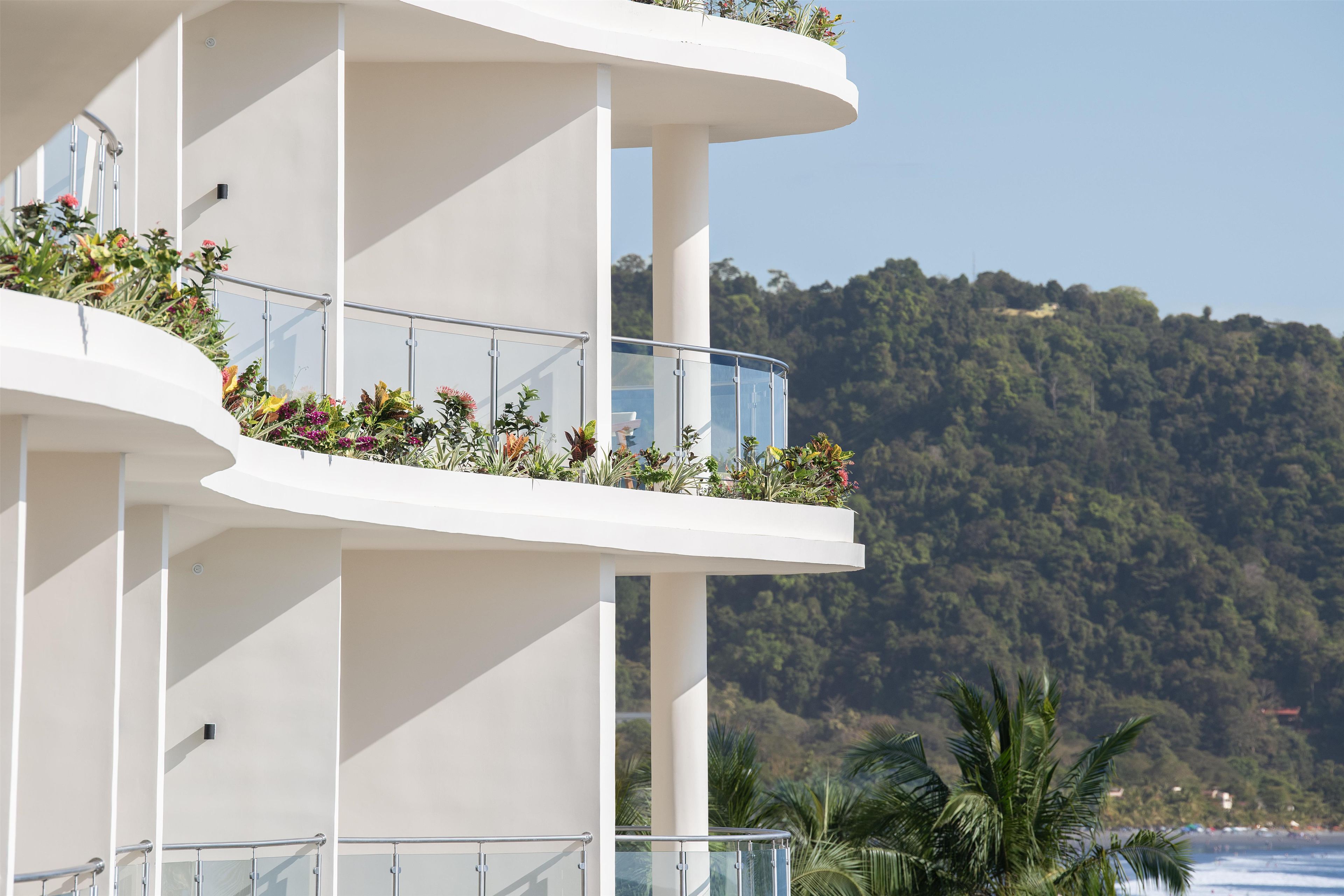 A multi-story hotel with balconies, each adorned with plants and offering views of a forested hillside.