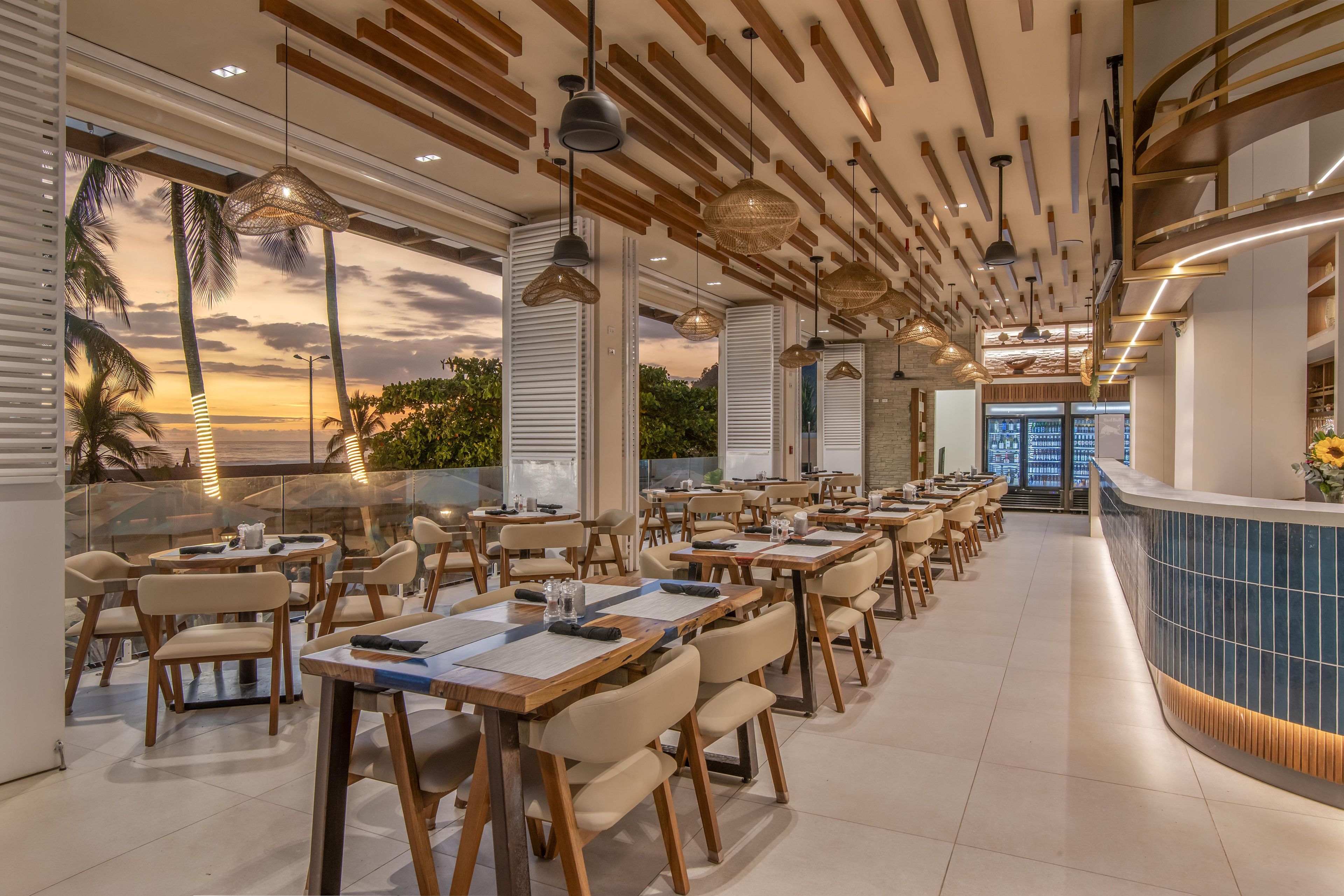 A restaurant with multiple tables and chairs, a curved counter, and a view of the sunset through large windows.