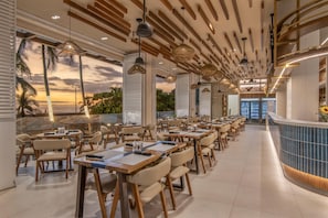 A restaurant with multiple tables and chairs, a curved counter, and a view of the sunset through large windows. - Clarita's Beach Bar & Resort, WorldHotels Elite (Jacó)