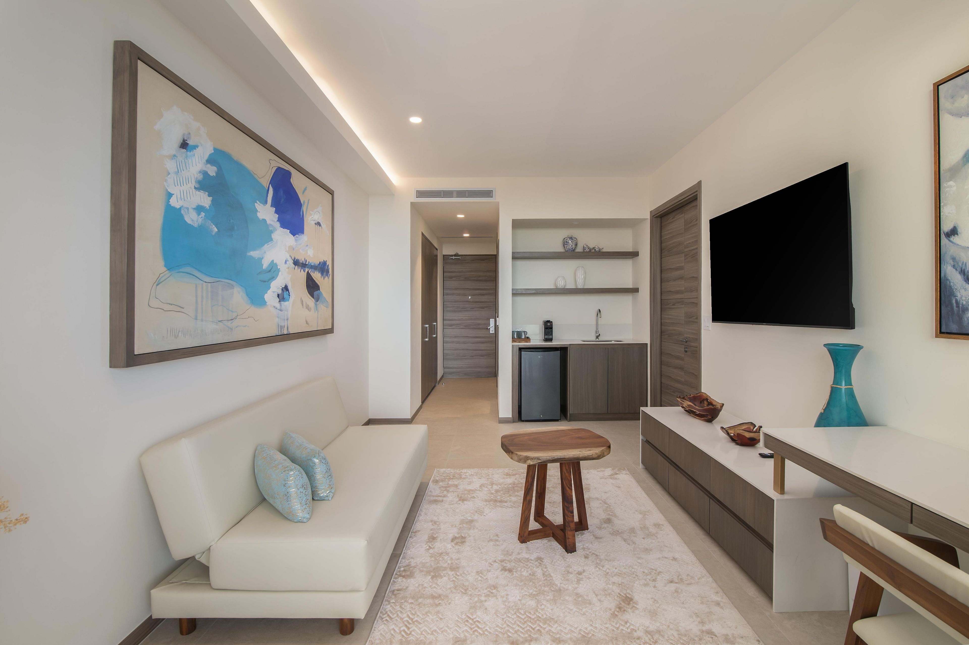 A modern living room with a sofa, a coffee table, a TV, and a kitchen area with a fridge and shelves.