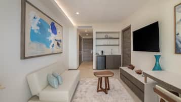 Una sala de estar moderna con sofá, una mesa de centro, televisor y una zona de cocina con refrigerador y estanterías.